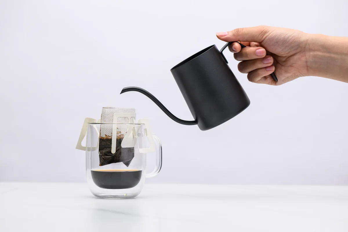 Hot water being poured from a black gooseneck kettle into a Sampan drip bag resting on a g