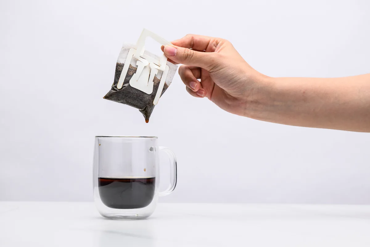 A hand lifting the spent Sampan drip bag away from a glass mug of finished coffee
