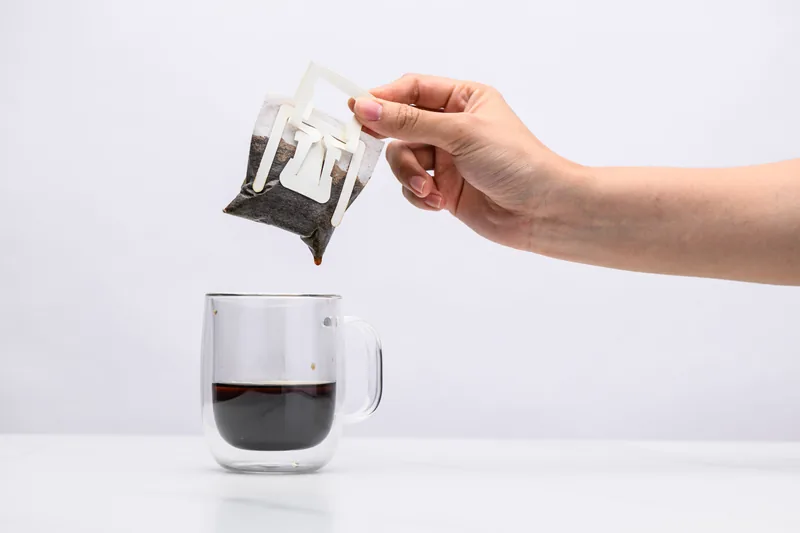 A hand lifting the spent Sampan drip bag away from a glass mug of finished coffee